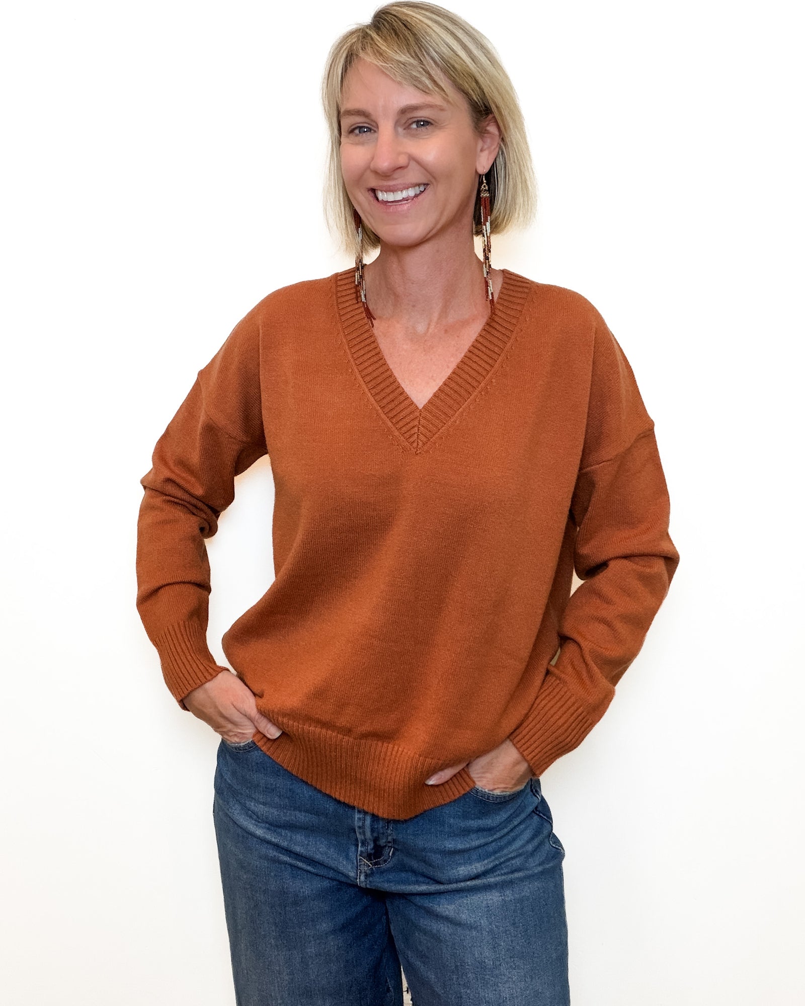 A person wearing a rust-orange ribbed V-neck sweater with long sleeves, paired with blue jeans, shown against a plain white background.