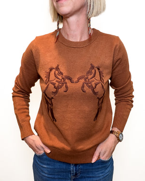 A woman wearing a rust-brown crewneck sweater with a dark brown embroidered design of two horses facing each other on the chest, paired with blue jeans against a plain white background.