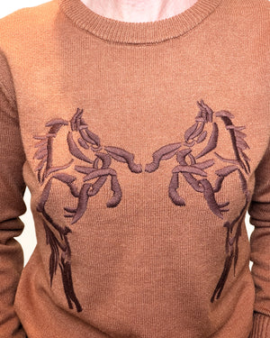 A close-up of a ribbed knit crewneck sweater in a warm camel brown color, featuring a symmetrical embroidered design of two stylized horses facing each other across the chest.