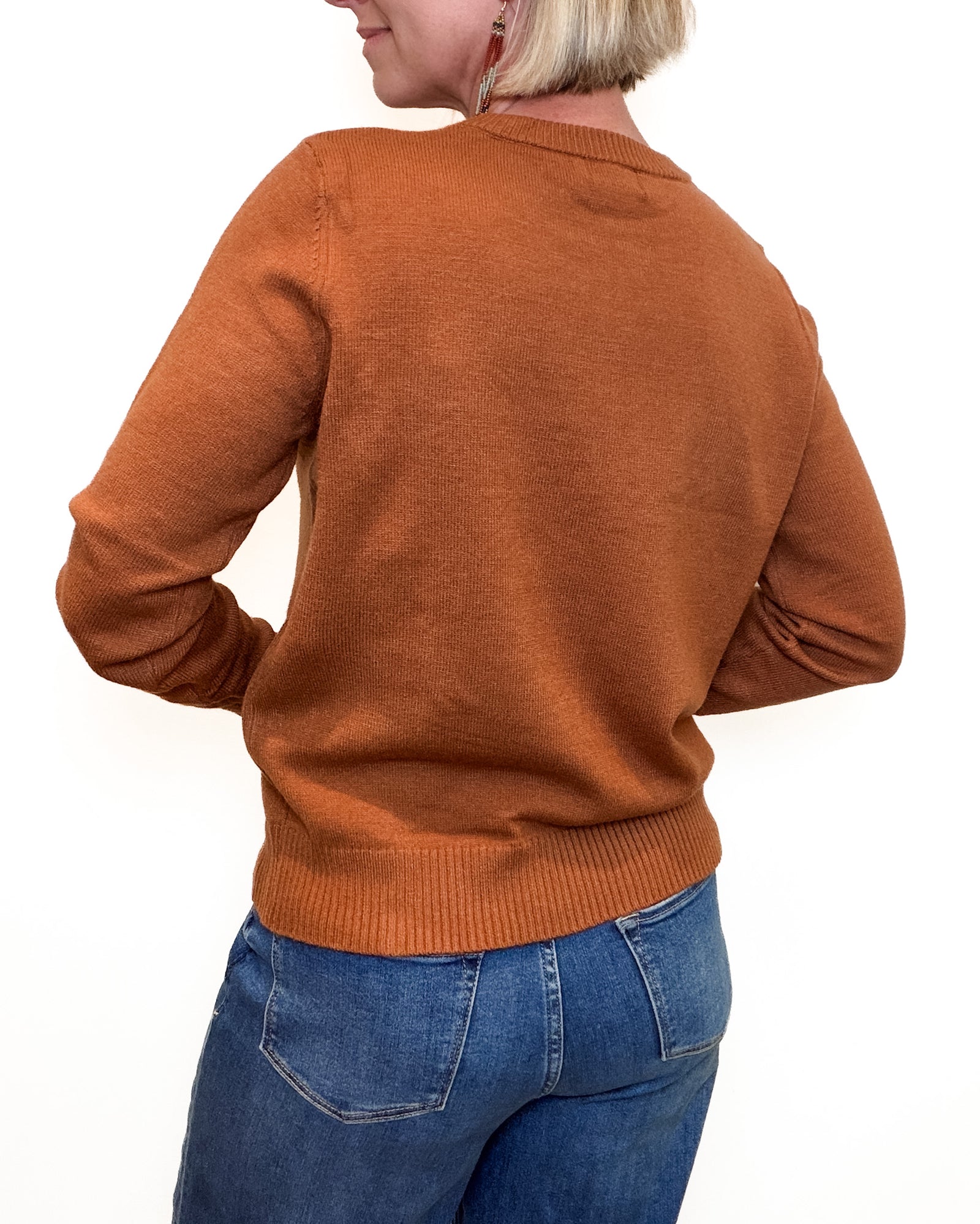 A woman wearing a rust-brown crewneck sweater with a dark brown embroidered motif of two horses facing each other on the chest, paired with blue jeans against a plain white background.