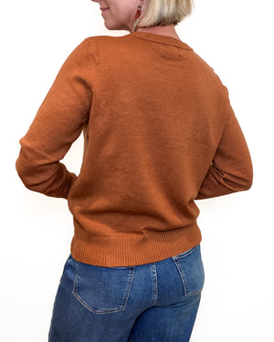 A woman shown from the back wearing a rust-orange crewneck sweater and blue jeans against a plain white background.