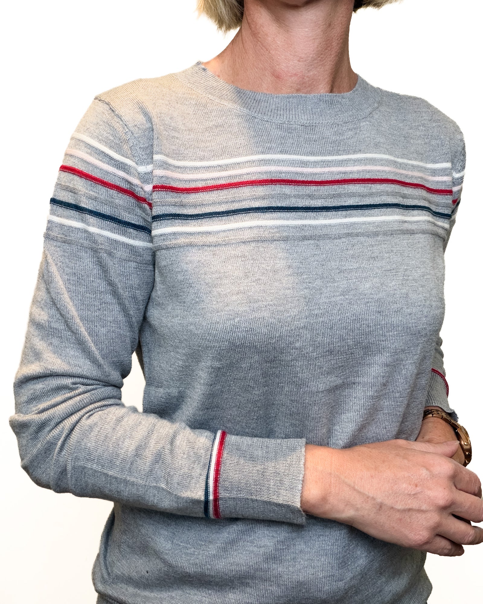 A person wearing a heather gray crewneck sweater with thin horizontal stripes in red, white, and navy across the chest and cuffs, paired with blue jeans against a plain white background.