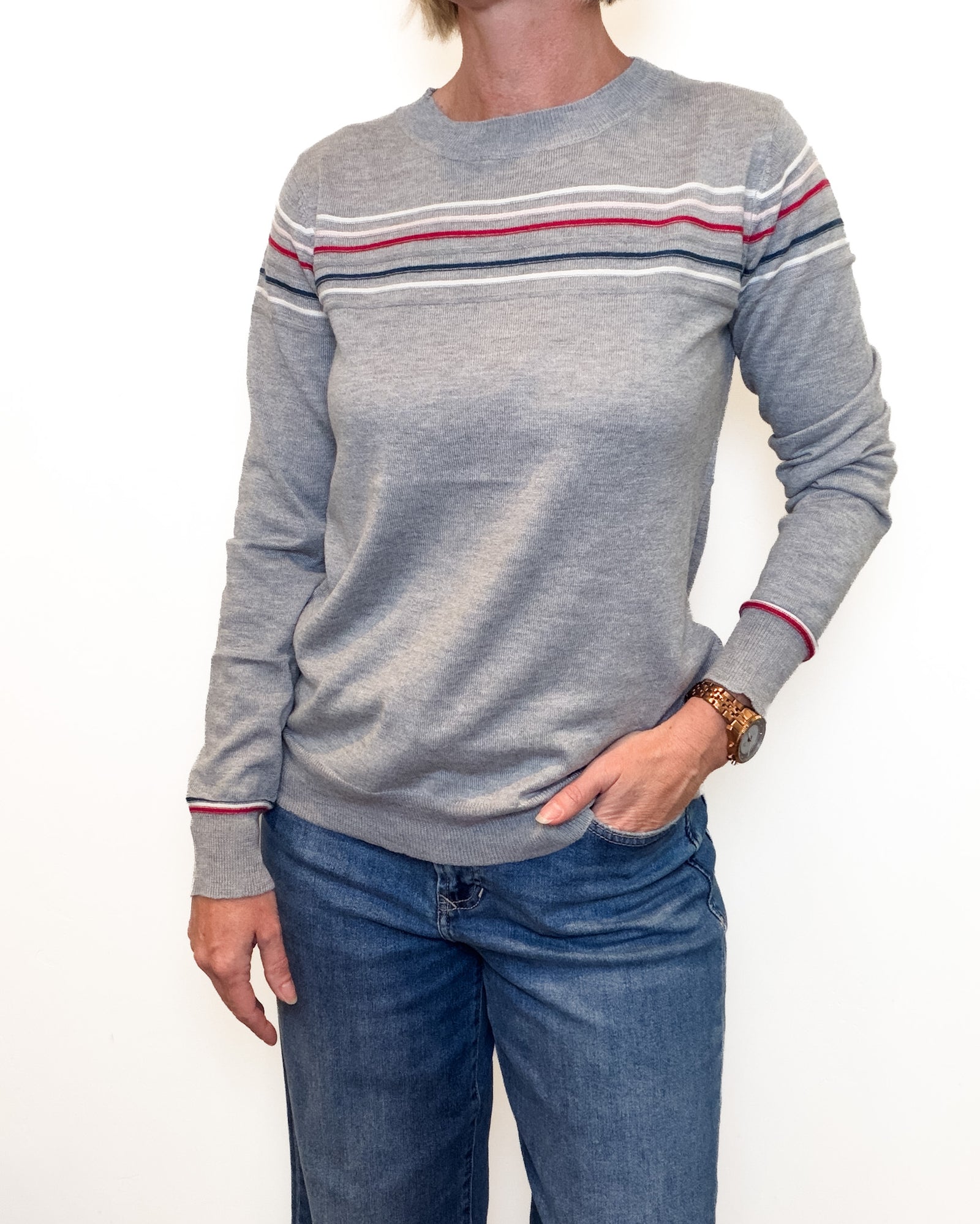 A person wearing a heather gray crewneck sweater with thin horizontal stripes in red, white, and navy across the chest and cuffs, paired with blue jeans against a plain white background.