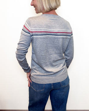 A woman shown from the back wearing a heather gray long-sleeve sweater with thin horizontal stripes in red, white, and navy across the shoulders and upper back, paired with blue jeans against a plain white wall.