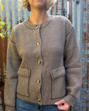 A woman wearing a ribbed knit cardigan in taupe brown with gold-tone buttons and two front flap pockets, paired with blue jeans, standing against a rustic corrugated metal wall.