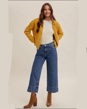 A woman modeling a mustard yellow knit cardigan over a white ribbed top, paired with blue wide-leg cropped jeans and brown ankle boots, against a plain beige studio background.