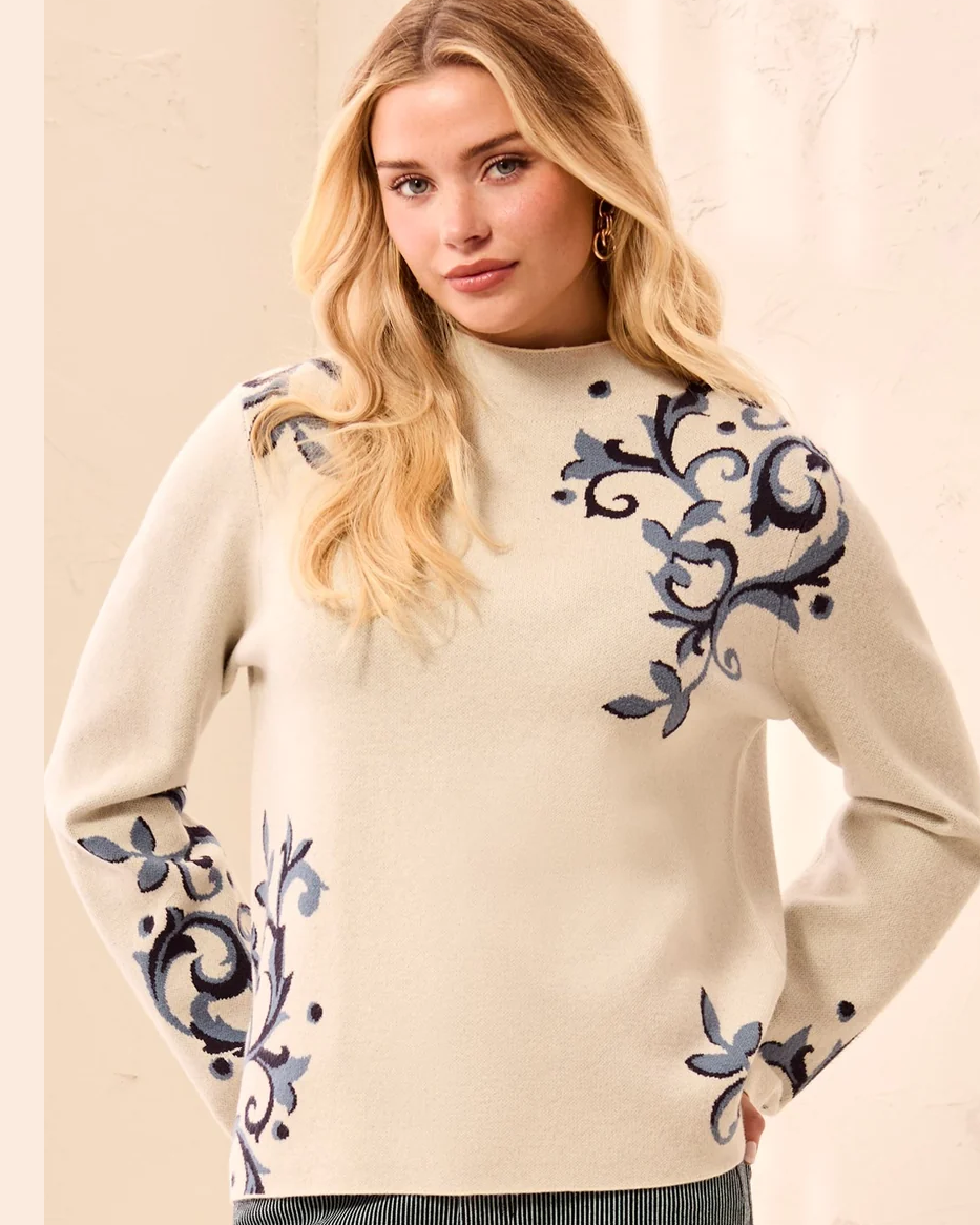 A woman modeling a cream knit sweater with blue floral embroidery on the sleeves and hem, paired with dark gray wide-leg jeans and beige sneakers in a bright indoor setting.