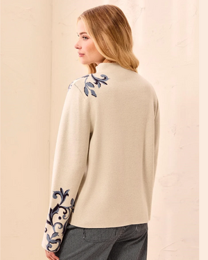 A woman shown from the back wearing a cream-colored knit sweater with blue floral embroidery on the shoulders and sleeve cuffs, paired with gray trousers, set against a neutral beige background.