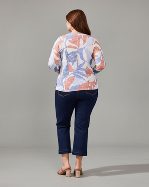 Woman wearing a colorful floral-patterned top and dark blue jeans in a room with a white wall and plant.