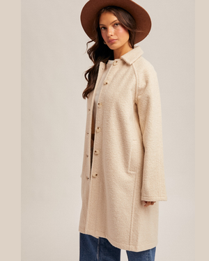 A woman wearing a long, light beige textured coat with brown buttons, paired with a brown wide-brim hat and blue jeans, standing against a neutral beige background.