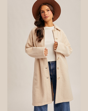A woman wearing a long, oversized beige knit coat over a white top and blue jeans, styled with a brown wide-brim hat against a plain light beige studio background.