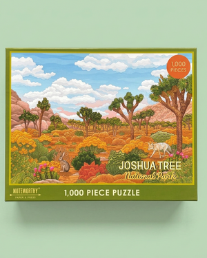 A 1,000-piece jigsaw puzzle box featuring an illustrated landscape of Joshua Tree National Park with blue sky, white clouds, green trees, and warm orange and yellow brush. The box has a green border and reads 'Joshua Tree National Park' and '1,000 Piece Puzzle'.