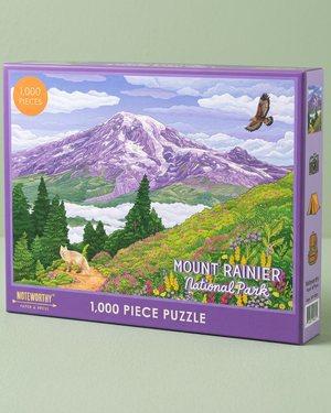 A 1,000-piece jigsaw puzzle box featuring an illustrated mountain landscape labeled Mount Rainier National Park. The box has a purple border and shows a colorful scene with a purple mountain, green hills, trees, clouds, and a bird.