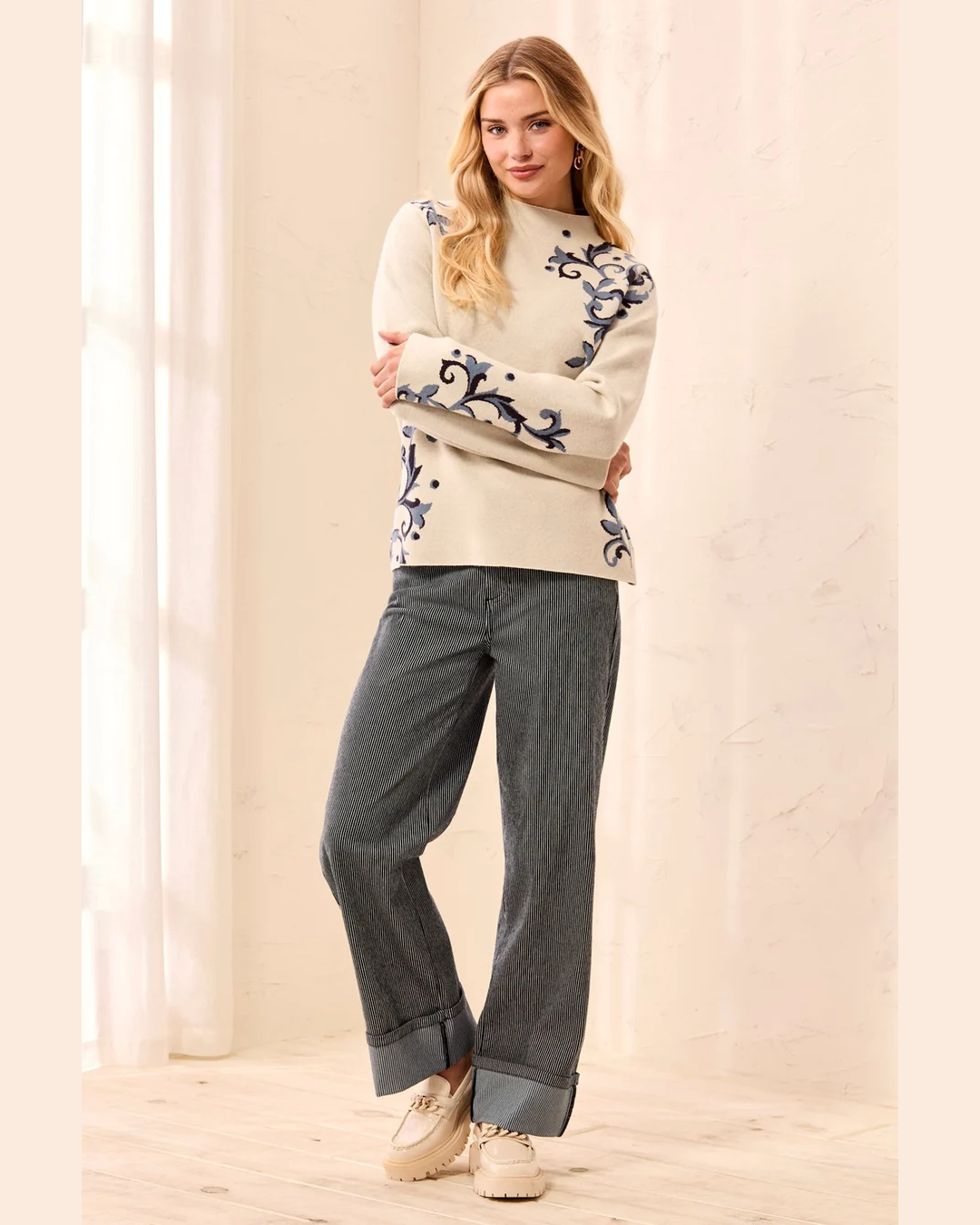 A woman modeling a cream knit sweater with blue floral embroidery on the sleeves and hem, paired with dark gray wide-leg jeans and beige sneakers in a bright indoor setting.
