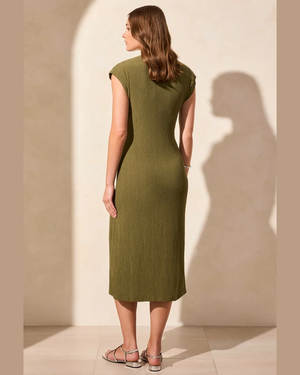 A woman stands with her back to the camera wearing a fitted olive green midi dress with cap sleeves and silver strappy heels, set against a warm beige wall with a soft shadow.