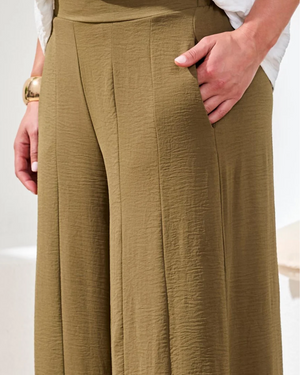 A close-up of a person wearing high-waisted, wide-leg trousers in a textured olive-brown fabric. The pants feature a high waistband, front pleats, and side pockets, with the model's hand resting in one of the pockets.