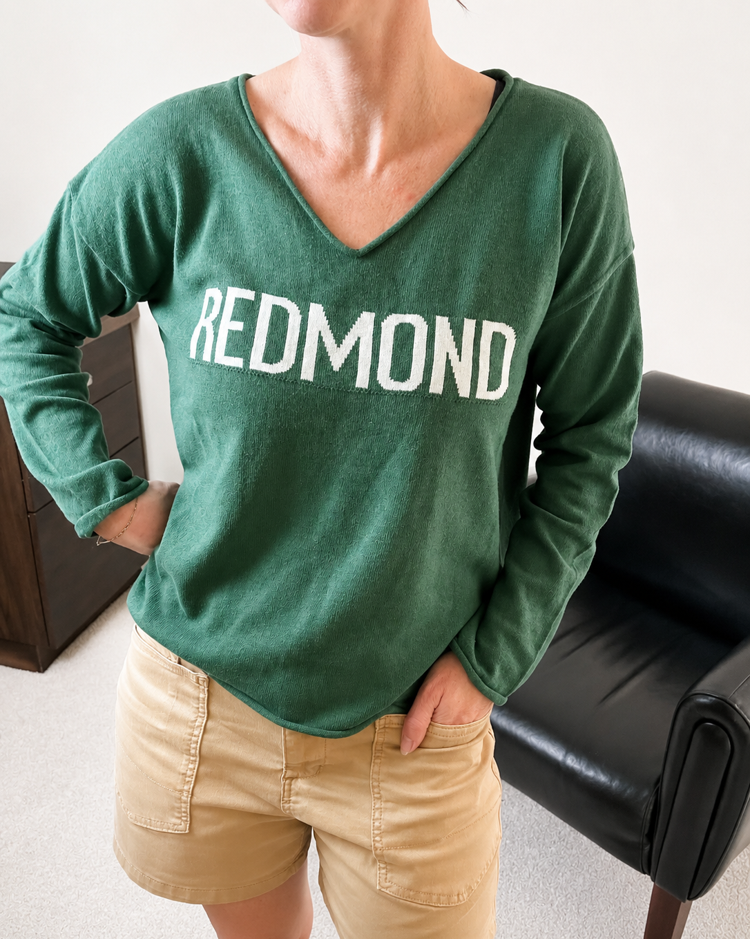 A person wearing a green long-sleeve V-neck sweatshirt with 'REDMOND' printed in white across the chest, paired with tan shorts, standing indoors near a black chair.