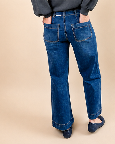 A person wearing blue wide-leg denim jeans with a dark gray top and navy flats, standing against a solid beige studio background.