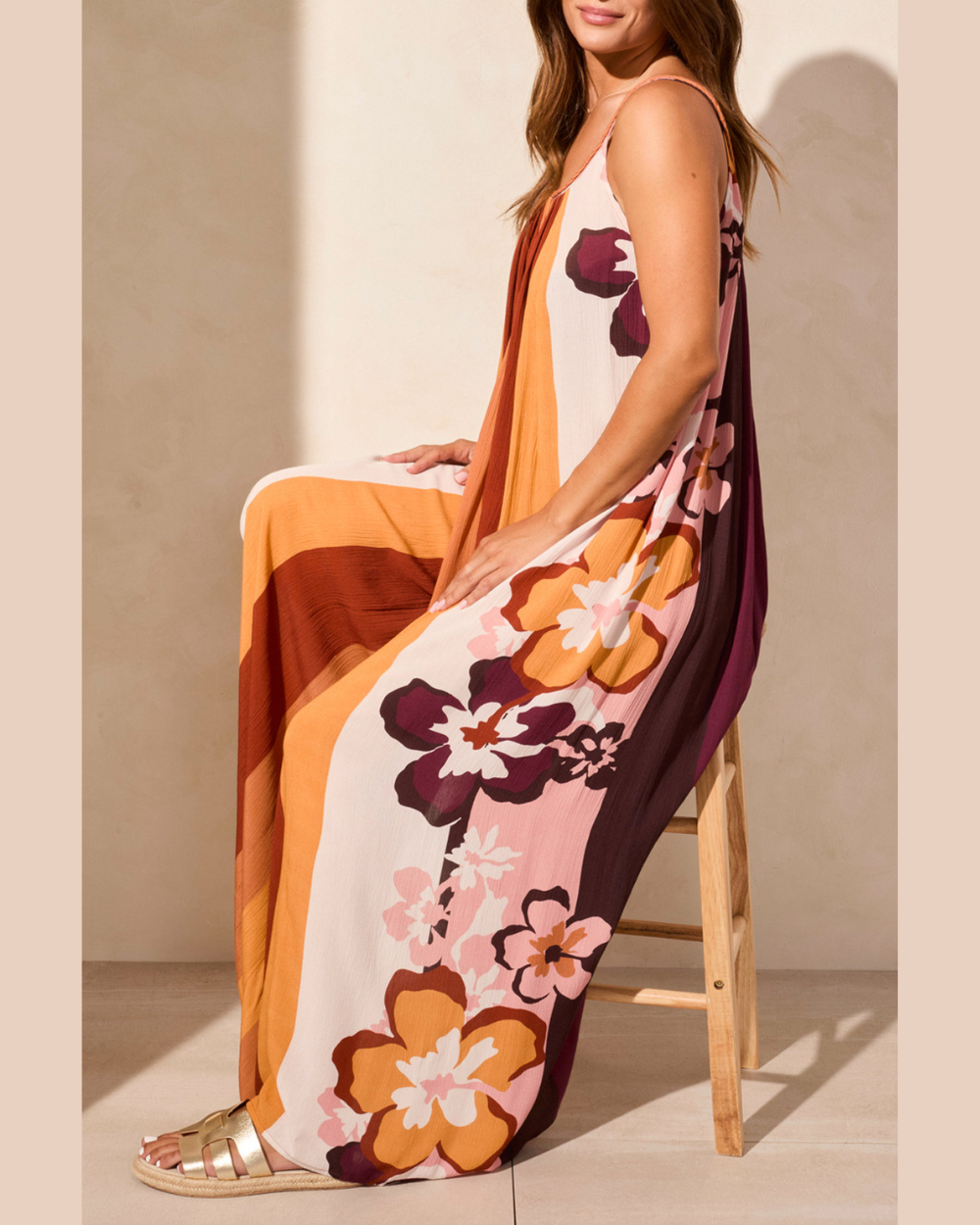 Woman wearing a floral dress with orange, pink, and brown colors sitting on a wooden stool.