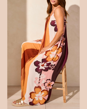Woman wearing a floral dress with orange, pink, and brown colors sitting on a wooden stool.