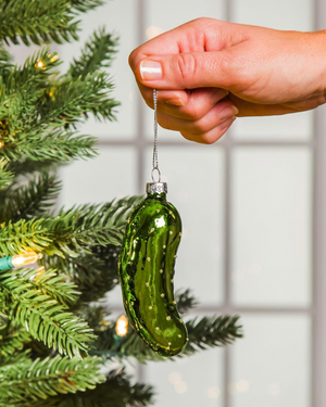 Green pickle ornament being held by a hand against a Christmas tree background