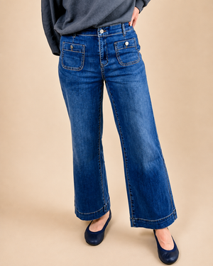 A person wearing blue wide-leg denim jeans with a dark gray top and navy flats, standing against a solid beige studio background.