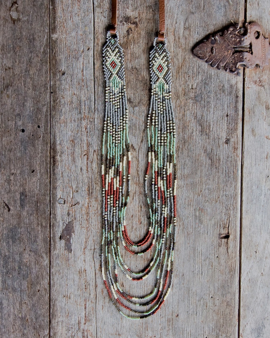 A multi-strand beaded necklace with a geometric pattern in turquoise, red, brown, and white, suspended from tan cords against a plain white background.