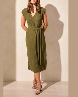 A woman models an olive green midi wrap dress with a V-neckline, cap sleeves, and a self-tie waist belt. She is wearing silver strappy high-heeled sandals and standing against a warm beige wall with soft shadows.