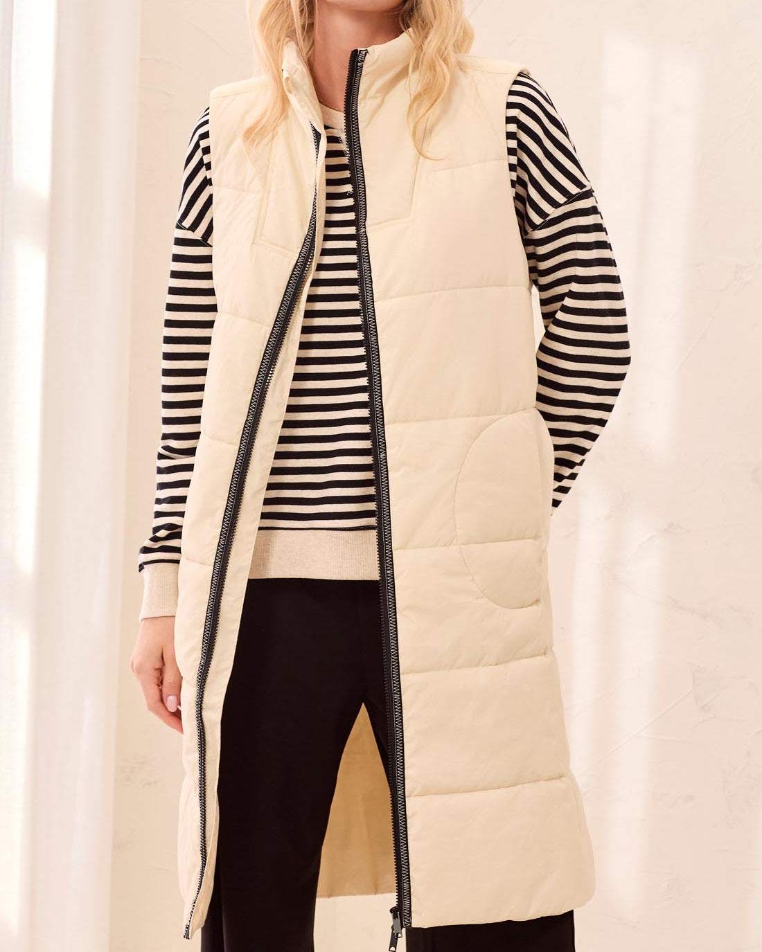 Woman wearing a long cream puffer vest over a striped shirt with a plain background