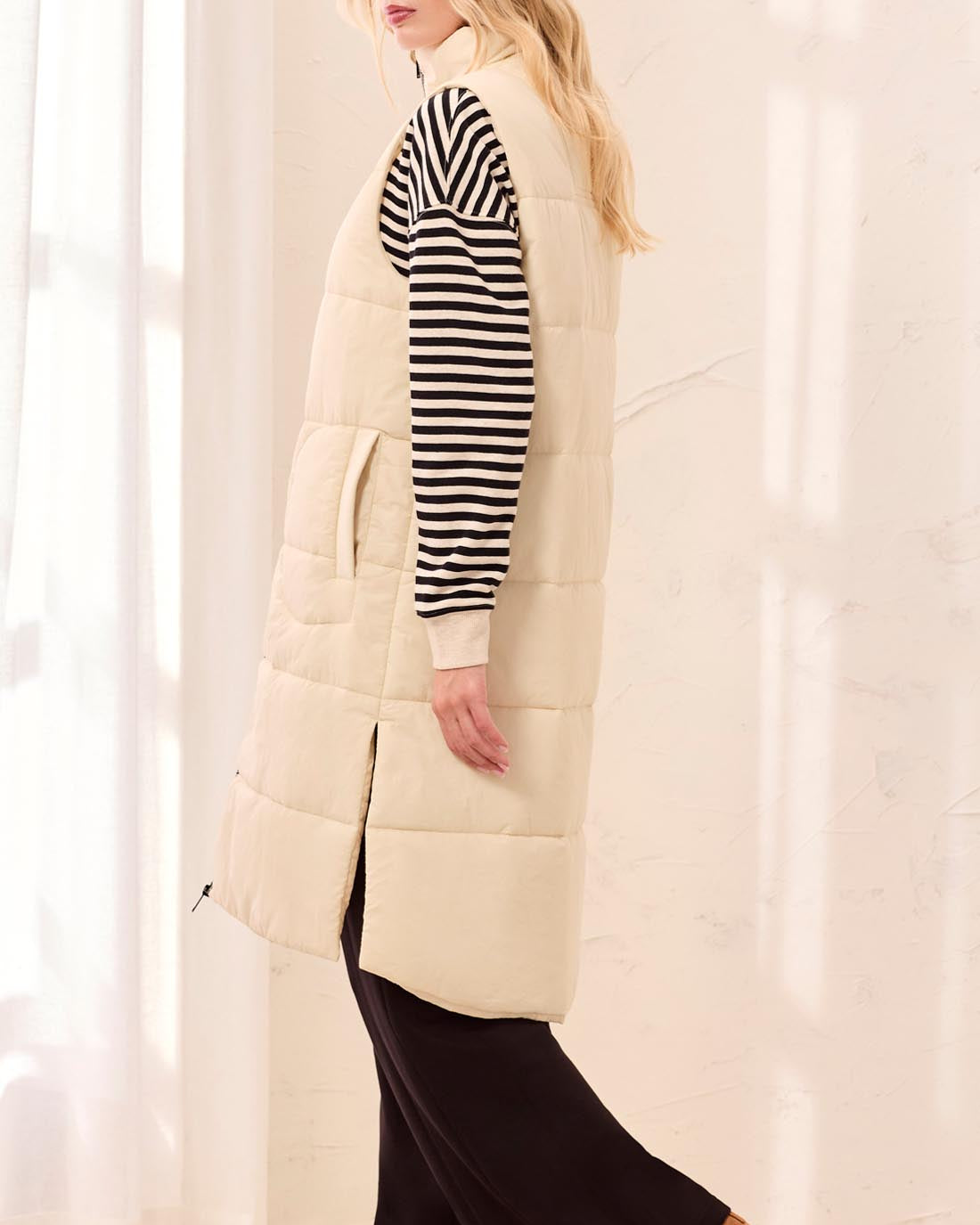 Woman wearing a long cream puffer vest over a striped shirt with a plain background