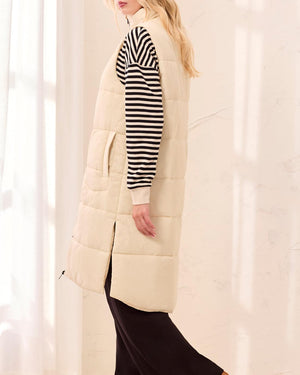 Person wearing a long cream puffer vest over a striped shirt with black pants and brown shoes on a white background