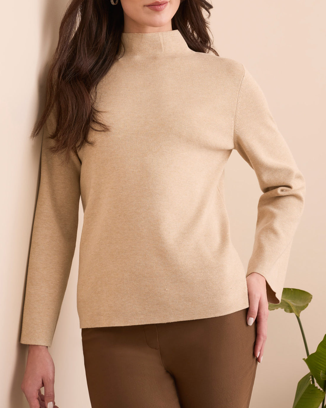 Woman wearing a beige sweater and brown pants against a neutral background