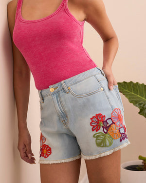 Person wearing a pink tank top and denim shorts with floral embroidery against a beige background