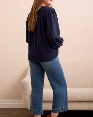 Person wearing a navy sweater, blue jeans, and brown boots standing in a room with a beige wall and wooden floor.