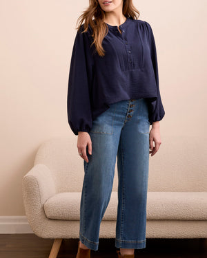 Woman wearing a navy blouse and blue jeans standing in a room with a beige sofa.