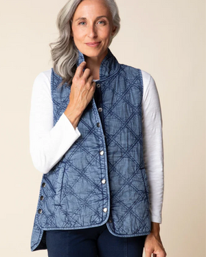 A woman with gray hair wearing a blue diamond-quilted vest over a white long-sleeve shirt and dark pants, standing against a plain beige background.