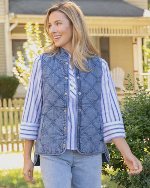 A woman wearing a blue quilted button-front vest over a blue and white striped shirt, standing outdoors in front of a house and garden.