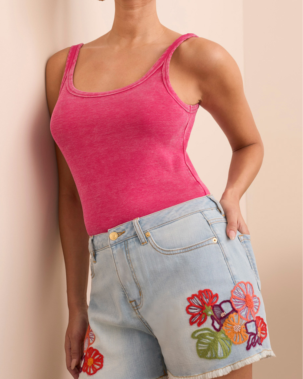 Person wearing a pink tank top and denim shorts with floral embroidery on a beige background